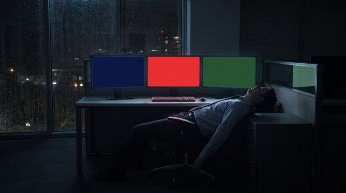 Operator asleep in a night office facing three solid-color monitors, a visual for an AI agent control tower with no on-screen text